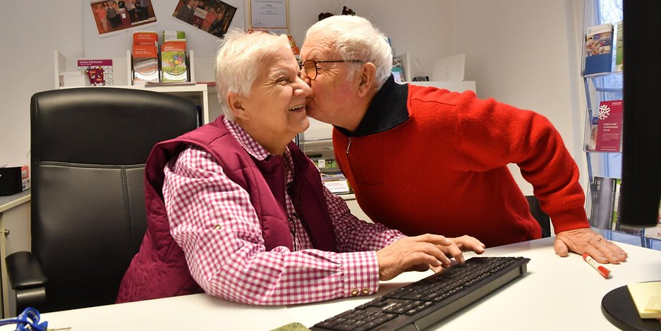 Eine ältere Frau sitzt am PC, ein älterer Herr beugt sich zu ihr und küsst ihre Wange: Ihre Ansprechpartner Marianne und Manfred Gabrowsky